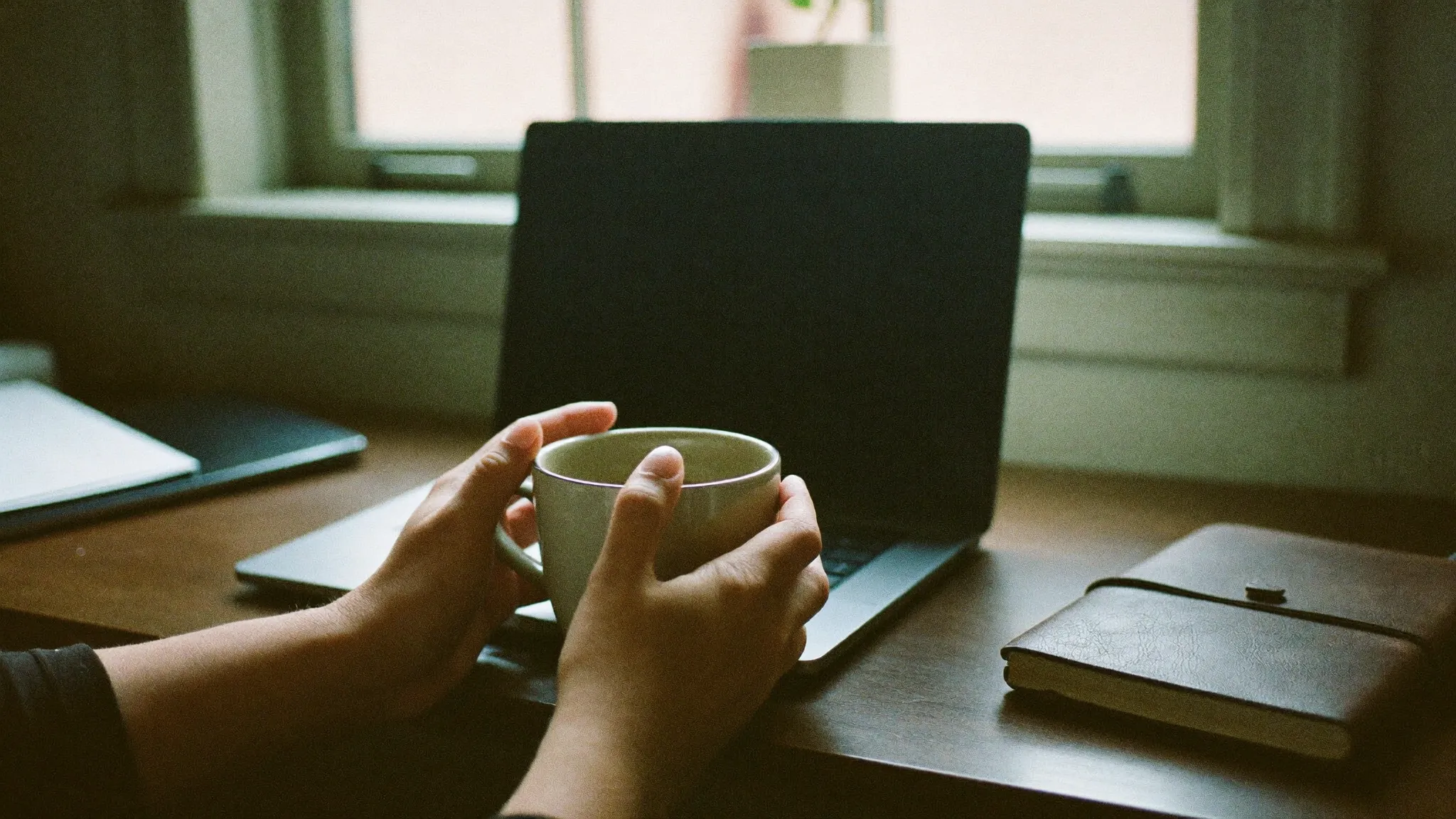 Hände halten eine warme Tasse am Fenster eines kleinen Arbeitszimmers, daneben ein zugeklappter Laptop