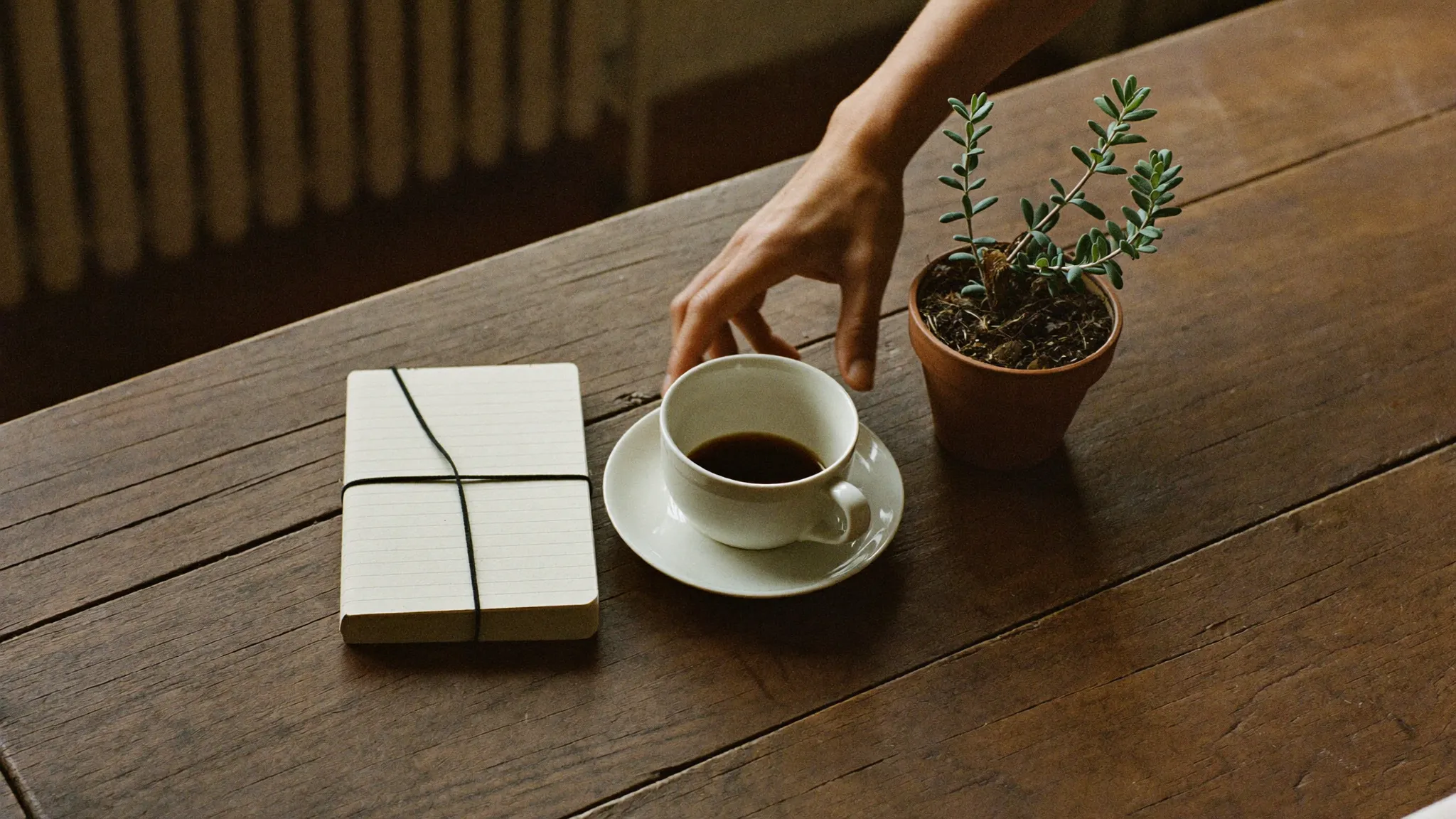 Hand stellt eine Tasse Kaffee neben ein geschlossenes Notizbuch auf einem rustikalen Holztisch