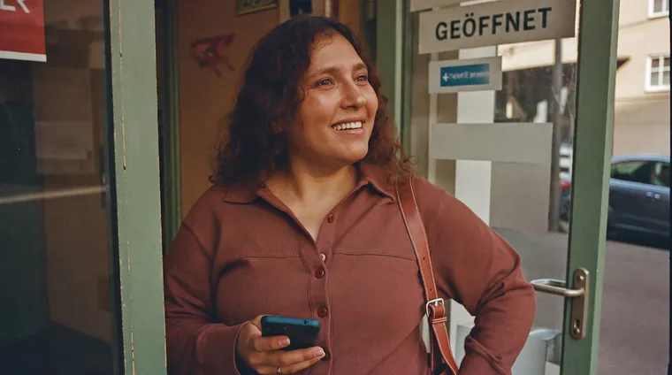 Händlerin steht in der Tür ihres Ladens und schaut mit dem Smartphone die Straße entlang